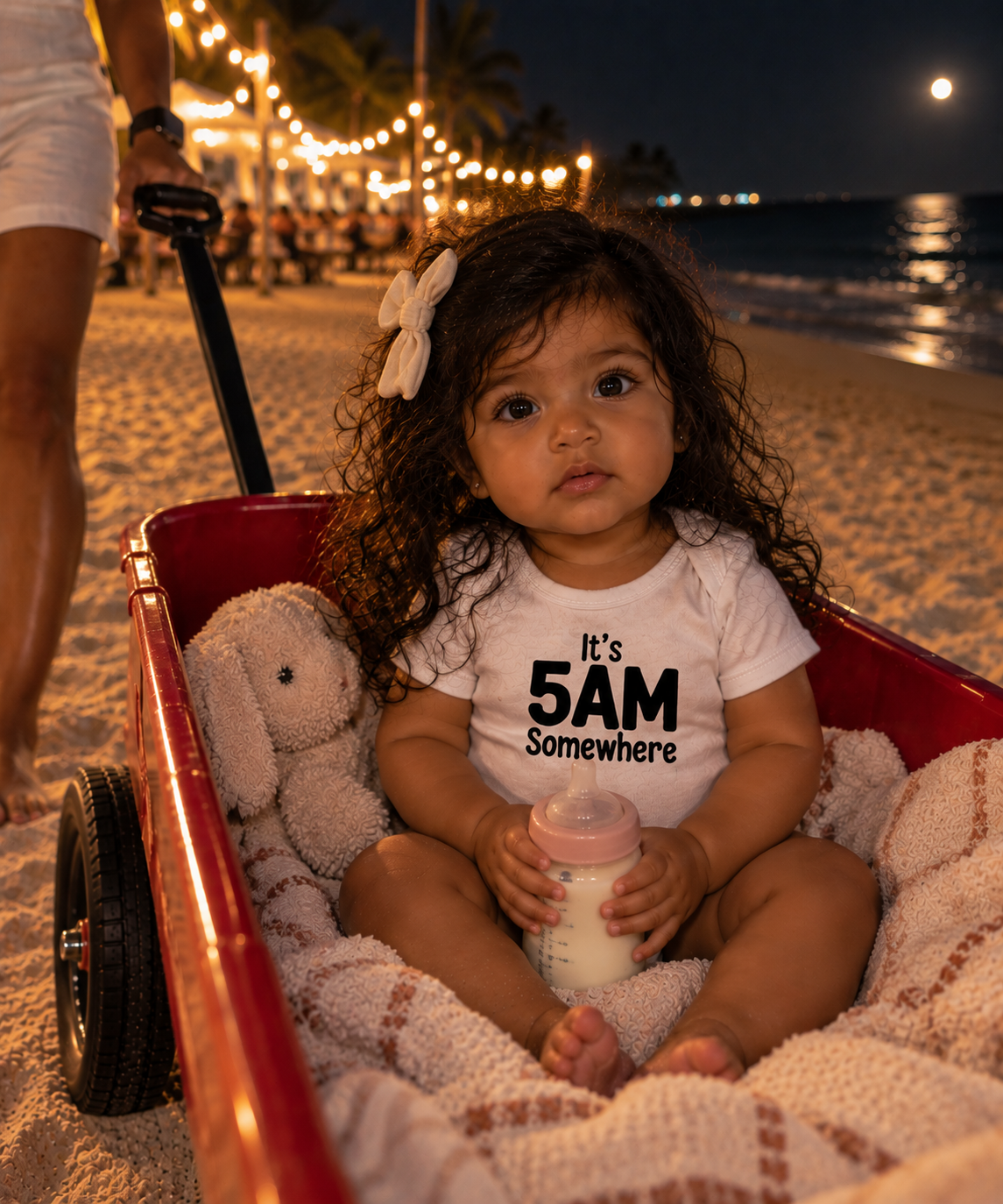 A toddler girl sits in a red wagon on a sandy beach at night, wearing a white graphic t-shirt that reads 'It's 5AM Somewhere' in black text. She holds a bottle and has a plush toy beside her.