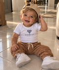 A smiling toddler sitting on a tiled floor indoors, wearing a white crewneck t-shirt with the 'Endless Threads Co.' logo, brown jogger pants, white sneakers, and a beige baseball cap.
