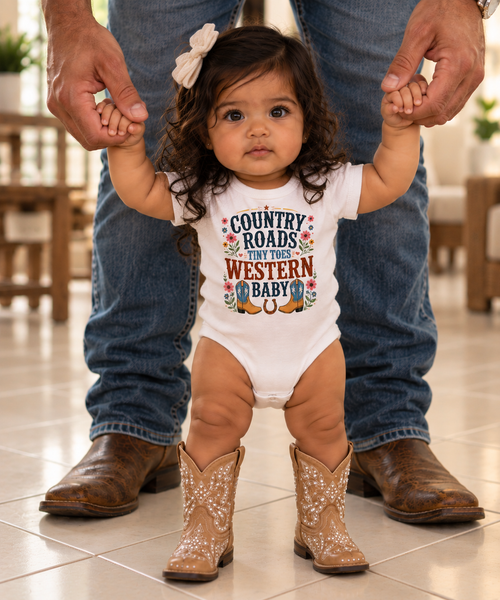 A toddler standing on a tiled floor, holding hands with an adult wearing blue jeans and brown cowboy boots. The child is wearing a white bodysuit with a colorful western-themed graphic that reads 'COUNTRY ROADS LEAVE THE WESTERN BABY ALONE' and tan cowboy boot prints on the legs.