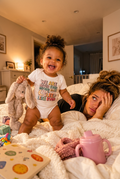 A baby wearing a white graphic t-shirt with the text 'WE ARE NEVER EVER GETTING BORED SLEEP LIKE EVER' stands on a white textured blanket next to a woman in bed. A pink sippy cup is in the foreground, and a stuffed bunny is held by the baby.