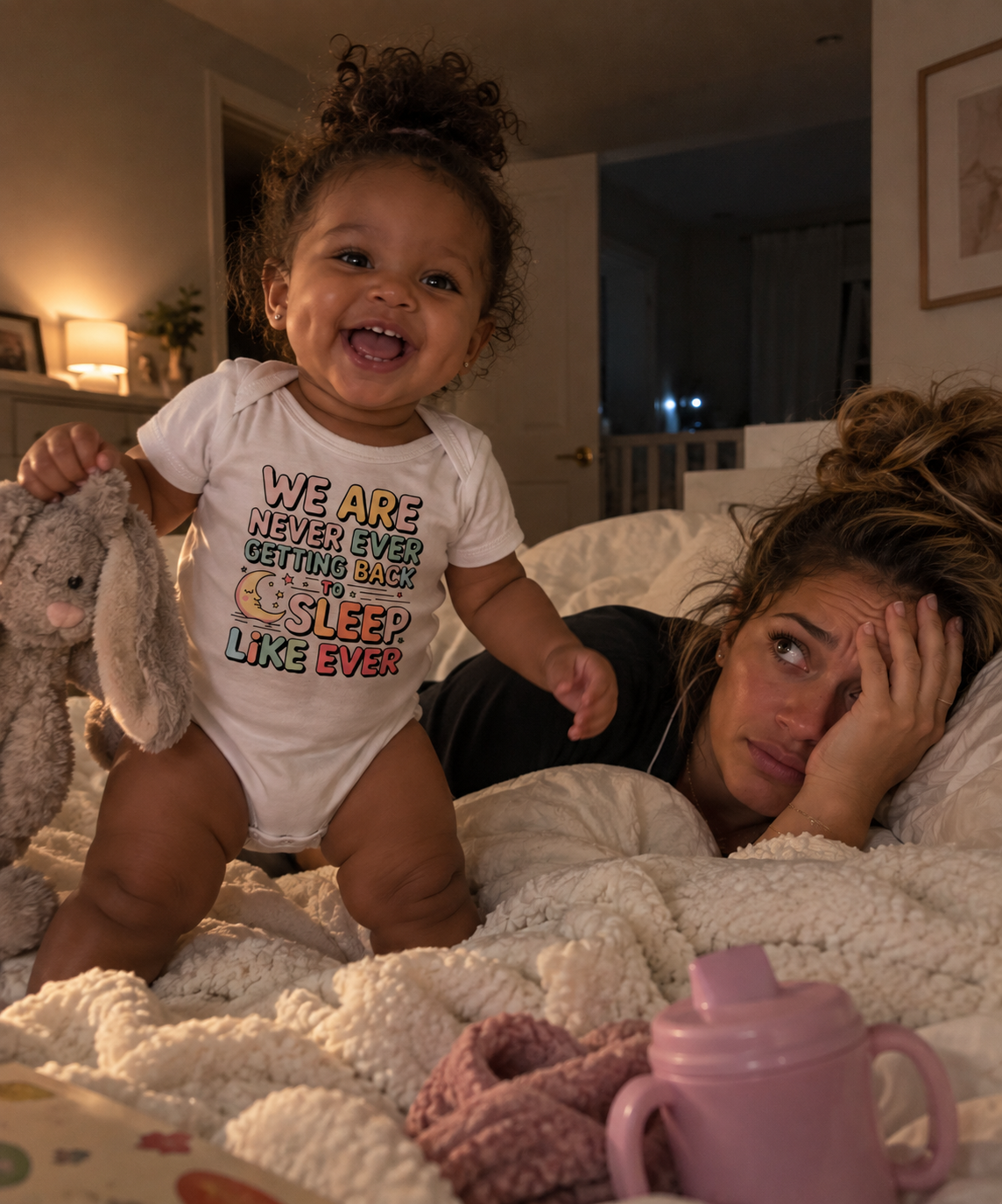 A baby wearing a white graphic t-shirt with the text 'WE ARE NEVER EVER GETTING BORED SLEEP LIKE EVER' stands on a white textured blanket next to a woman in bed. A pink sippy cup is in the foreground, and a stuffed bunny is held by the baby.