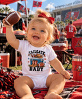 A baby girl sitting outdoors at a tailgate event wearing a white short-sleeve bodysuit with a Tampa Bay Buccaneers graphic that reads 'TAILGATE BABY'. She is holding a football up in one hand, with a red hair bow, red cooler, and stadium background visible.